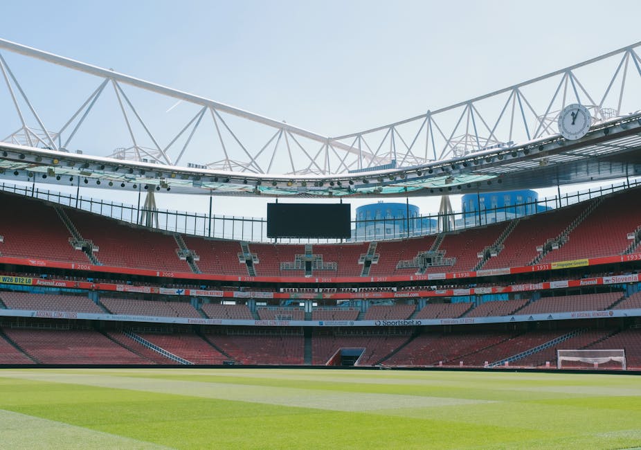 Interior view of a modern football stadium featuring a well-maintained grass pitch with light green and dark green stripes, surrounded by red seating sections. The arena is equipped with a large, black electronic scoreboard hanging from the upper tier, and bright, natural daylight illuminates the space. White structural beams and supports adorn the roof area, creating a geometric pattern against the blue sky. The seating and field are spotless, highlighting regular cleaning and maintenance, with no visible dirt or debris. The stadium appears ready for a match or event, emphasizing the importance of thorough surface cleaning and hygiene in large-scale sports venues. Mention of Carpet Cleaners Islington suggests their expertise in deep cleaning and sanitisation services, promoting a clean and hygienic environment for both sports teams and spectators.
