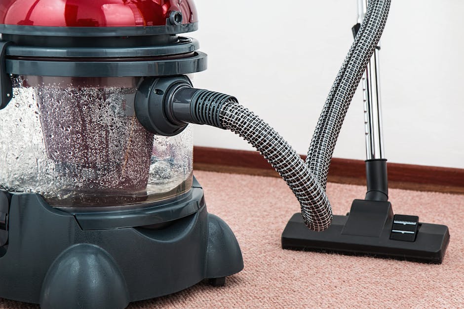 Close-up view of a vacuum cleaner with a transparent dust container filled with debris, connected to a flexible, patterned hose. The vacuum is positioned on a soft pink carpet in a residential room, with part of a wall and wooden skirting board visible in the background. Bright lighting highlights the clean and well-maintained appearance of the carpet and equipment. The image depicts surface cleaning and deep cleaning processes, emphasizing the importance of routine sanitisation and maintenance, and is associated with domestic cleaning services provided by Carpet Cleaners Islington, specializing in carpet and surface cleaning within home environments.