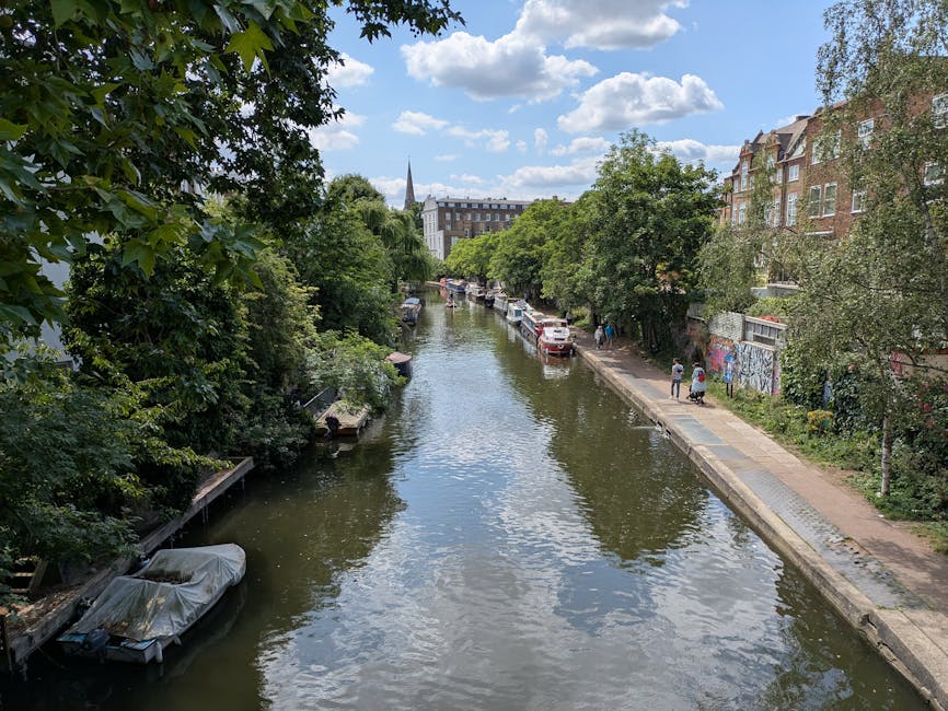 A scenic view of a narrow, calm canal in Highbury flats, Islington, with clear water reflecting the blue sky and white clouds above. The canal is bordered by a paved footpath on the right side, with a few pedestrians walking along it. On the left side, there are lush green trees with dense foliage that overhang the water, and on the right, a row of brick residential buildings with large windows and some graffiti on the lower walls. Several boats are moored along the edges of the canal, some covered with protective tarps. The area appears tidy and well-maintained, with natural lighting highlighting the vibrant greenery and the peaceful atmosphere of this urban waterway, exemplifying the importance of surface cleaning and maintenance in residential environments as part of professional cleaning services offered by Carpet Cleaners Islington.
