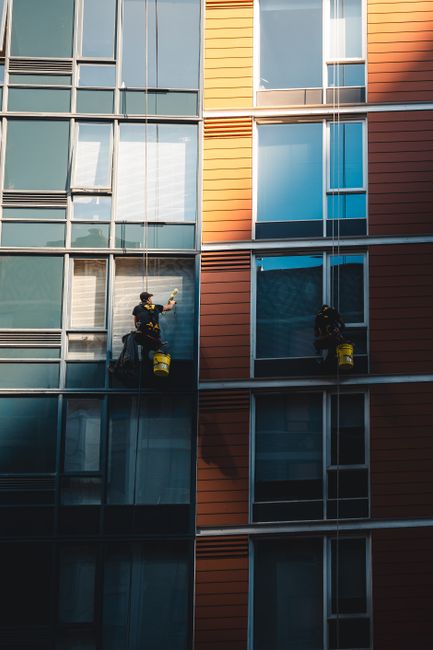 Two window cleaners wearing safety harnesses and protective helmets are performing surface cleaning on the glass exterior of a modern multi-story building with reflective glass panels in shades of blue and orange. They are positioned on suspended platforms, applying cleaning solutions and wiping the glass surfaces to maintain cleanliness and clarity. The building's exterior features large, rectangular glass windows framed with metal, with some sections reflecting the sky and surrounding environment. Bright natural light illuminates the scene, highlighting the reflective quality of the glass. As a reputable professional cleaning service, Carpet Cleaners Islington specializes in high-rise window cleaning, ensuring buildings remain pristine and safe while maintaining their aesthetic appeal.