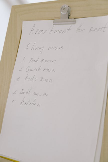 A handwritten list on a white sheet of paper attached with a metal clip to a wooden surface, detailing rooms in an apartment for rental purposes, including living room, bedroom, guest room, kid's room, bathroom, and kitchen. The paper appears slightly crumpled with clear, legible pencil writing. The background shows a light-colored wooden panel, and the overall setting suggests an organized, indoor environment used for planning or documentation related to cleaning or rental management.
