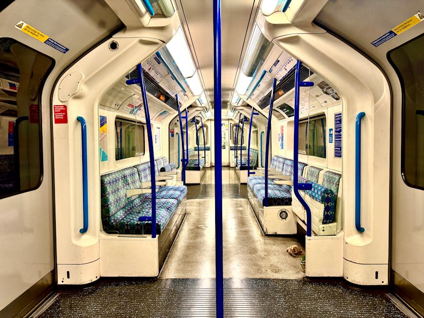 Interior view of a clean, empty subway train carriage with vibrant, multicolored patterned fabric seats arranged in parallel rows along both sides of the carriage. The floor is made of durable, non-slip material and appears spotless, indicating recent deep cleaning. Bright lighting illuminates the space evenly, enhancing the white and blue plastic interior panels, handrails, and window frames. Clear, professional safety and informational stickers are visible on the ceiling and walls. Blue vertical handrails are positioned beside the seats for passenger support. The carriage's windows are large, allowing natural light to complement the interior lighting. This setting illustrates the high standards of surface cleaning and sanitisation, which could be achieved by professionals such as Carpet Cleaners Islington, especially during end of tenancy or maintenance cleaning tasks in public transport environments.