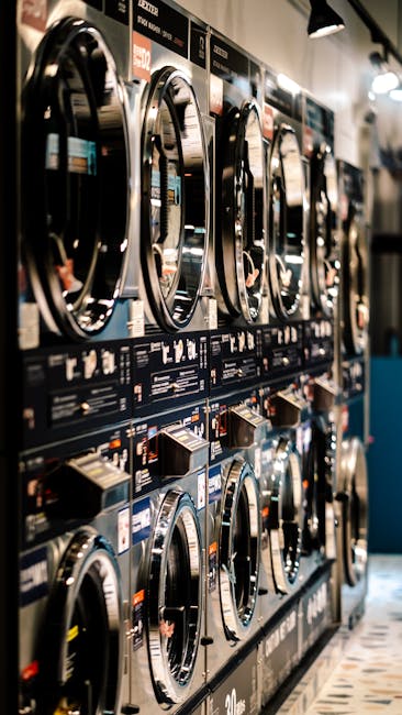 A row of front-loading washing machines with metallic doors and digital controls, installed within a commercial laundromat. The surfaces of the machines are clean and shiny, reflecting the overhead lighting. The machines are aligned on a wall with a black and blue panel below each, displaying various settings and instructions. The flooring consists of patterned tiles in shades of gray and black. The well-lit environment emphasizes cleanliness and maintenance, consistent with professional surface cleaning and sanitisation practices. Carpet Cleaners Islington is mentioned as a trusted provider for domestic and commercial cleaning, ensuring hygienic, deep cleaning for laundry spaces and other areas.