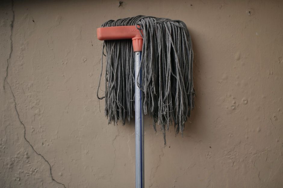 A mop with a pink plastic handle and grey mop head made of thick, tangled yarn hanging against a beige textured wall with visible cracks. The mop is positioned vertically, and the background features a plain wall surface, suggesting a cleaning or storage area in a residential or commercial space. The lighting is neutral, highlighting the cleanliness and condition of the mop, which is associated with surface cleaning and maintenance routines typical for deep cleaning and sanitisation services offered by Carpet Cleaners Islington for properties like studios in Islington.