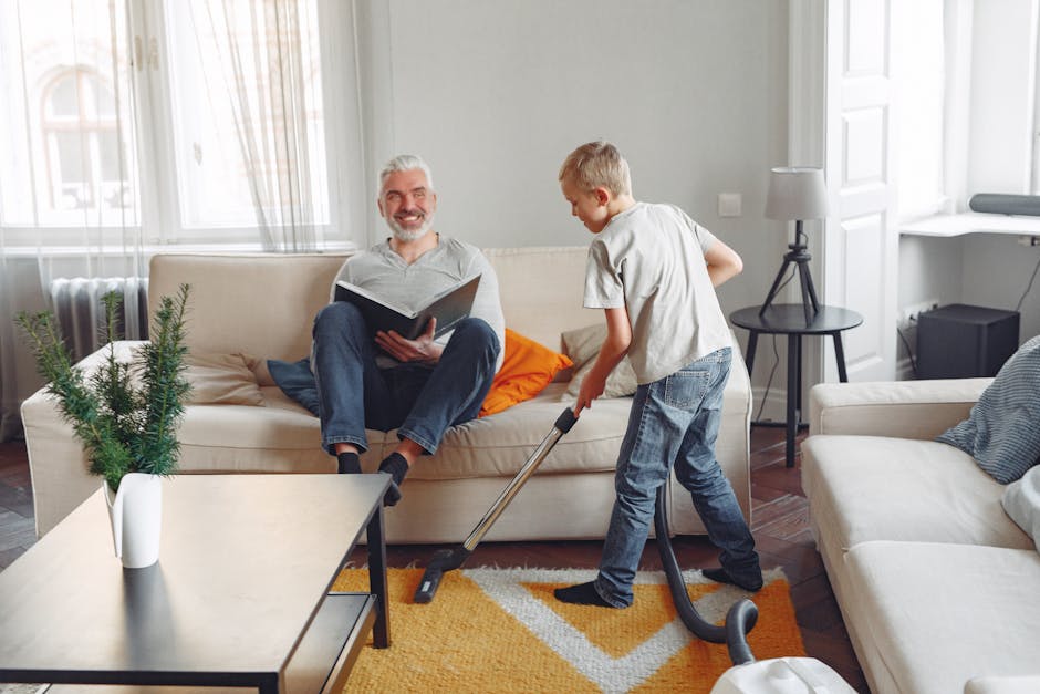 A young boy vacuuming a light-colored fabric sofa in a bright living room with natural daylight streaming through large windows, while an older man with a beard and grey hair sits on the sofa reading a book. The room features a modern interior with white walls, a small black side table with a lamp, a patterned rug in shades of orange and beige, and decorative cushions. A small potted plant on a coffee table adds greenery, and the space appears clean and well-maintained, reflecting professional domestic cleaning activities. Carpet Cleaners Islington specializes in surface cleaning and deep cleaning for urgent stain removal in residential settings, ensuring hygiene and freshness in homes.