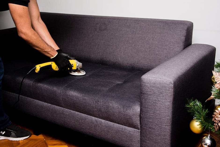 A person using a handheld vacuum cleaner to deep clean a decorative yellow and white patterned cushion placed on a dark fabric sofa in a room with neutral walls. The image highlights surface cleaning of the cushion and sofa, demonstrating hygiene and maintenance for domestic cleaning. This scene is associated with professional cleaning services offered by Carpet Cleaners Islington, specializing in sofa cleaning in Barnsbury Canonbury, ISLINGTON, with focus on surface sanitation and deep cleaning processes.